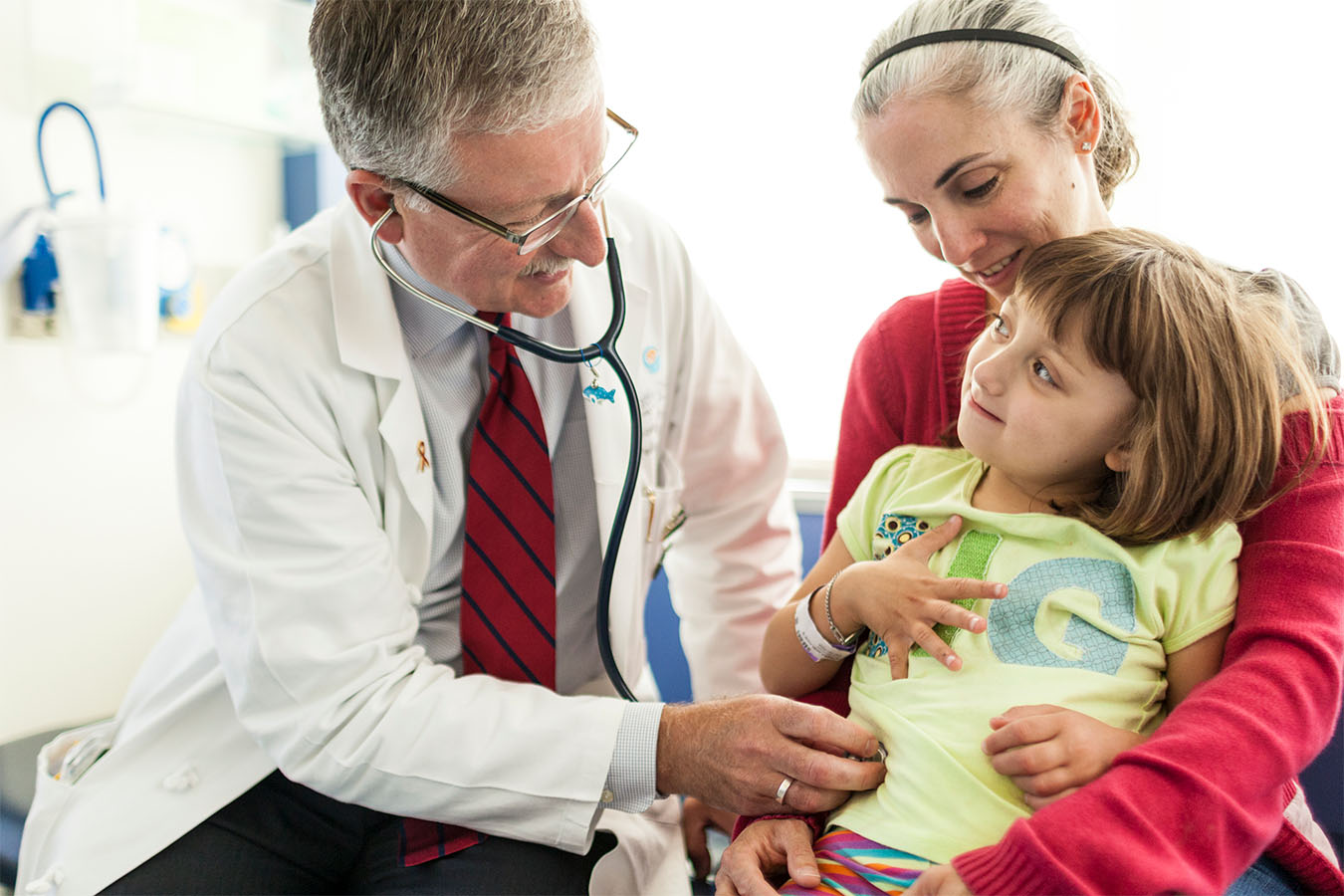 A doctor uses a stethoscope to listen to a child’s belly while a caregiver holds the child on their lap.