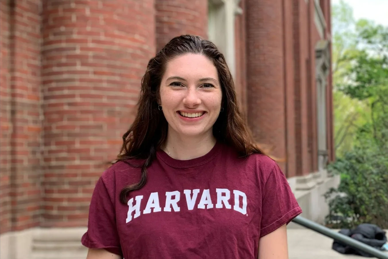A young woman stands in front of a brick building while wearing a Harvard sweatshirt