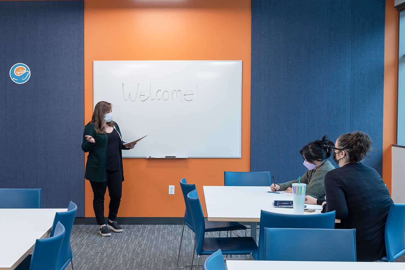 An insttructor stands in front of a whiteboard with the word "welcome" written on it.