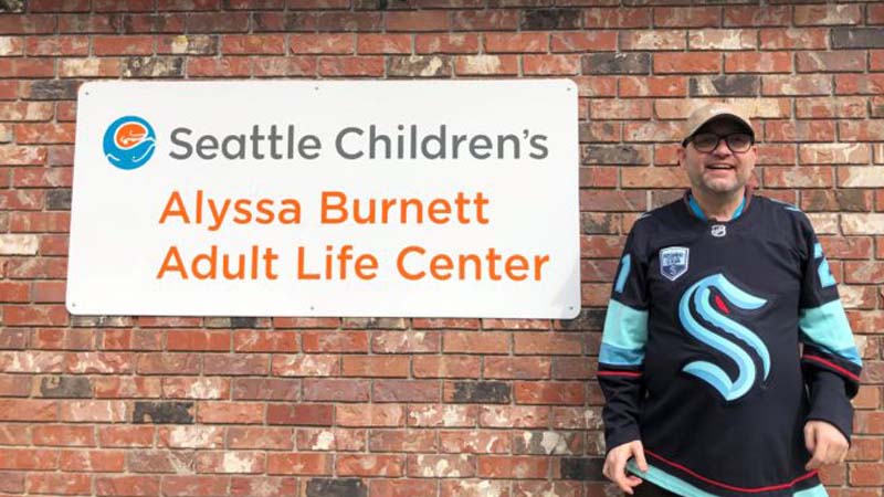 A man standing outside by a sign that says Seattle Children’s Alyssa Burnett Adult Life Center