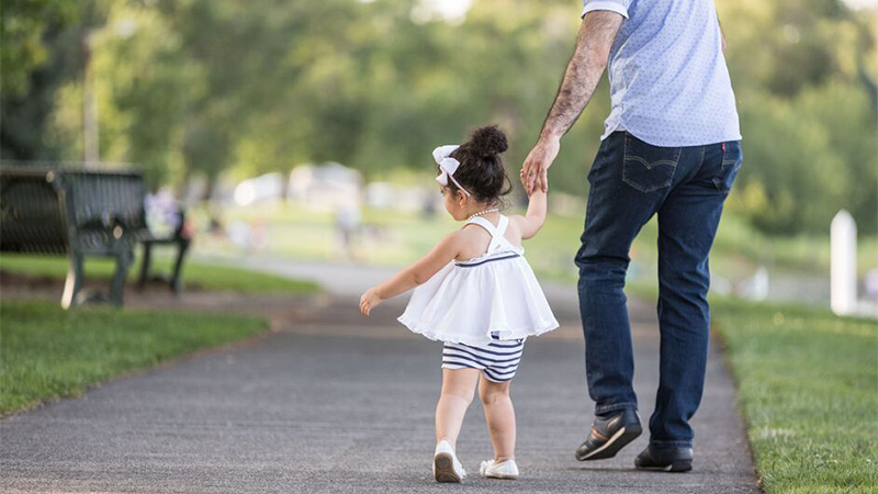 A father and young daughter walking down a sunny path hand in hand