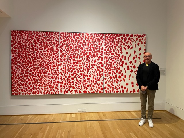 A man stands in front of a painting with red pieces on an ivory background