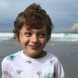 A young boy smiling while standing on a beach with large rocks in the waves behind him