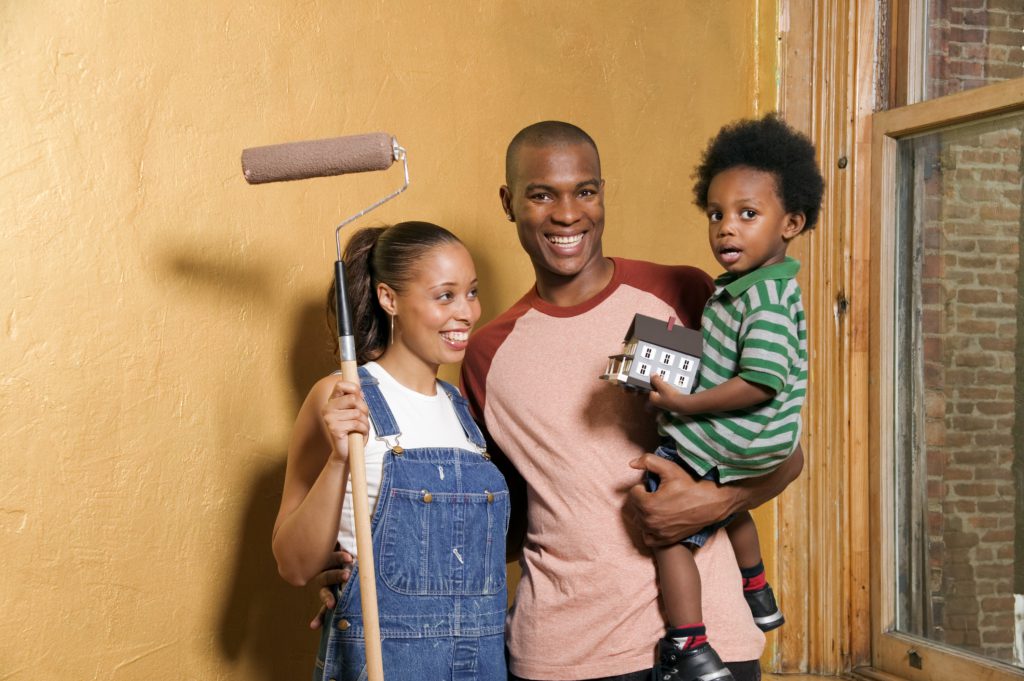 A mother and father, who is holding his son, post with a pain roller that is covered in the same color paint as the wall their standing in front of