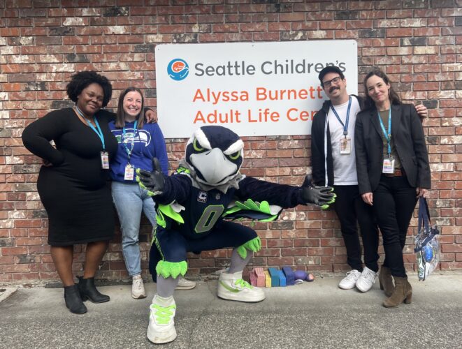 Blitz, the Seahawks mascot, poses with staff in front of the Seattle Children’s Alyssa Burnett Adult Life Center building