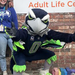 Blitz, the Seahawks mascot, poses with staff in front of the Seattle Children’s Alyssa Burnett Adult Life Center building