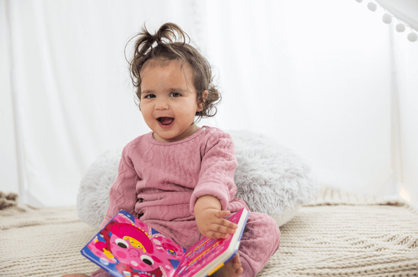 A little girl smiles at the camera while turning the page of a book