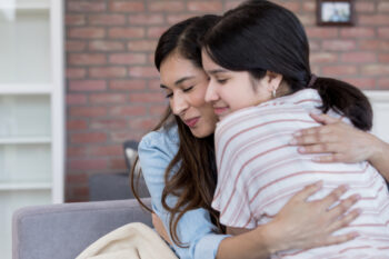 A mom and daughter embrace each other while sitting on a couch