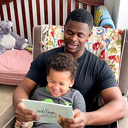 A son happily sits on his dad's lap in while he reads him a book