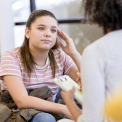 a teen girl sits on a couch facing a woman with a clipboard