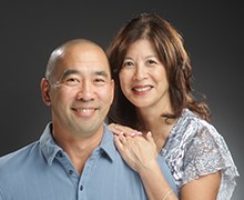 A middle-aged bald man is in a blue polo shirt and smiling straight towards the camera. His wife has medium length brown hair and is in a blue, white and grey patterned shirt. She rests her hands on his shoulder leaning into his side and also smiles towards the camera.