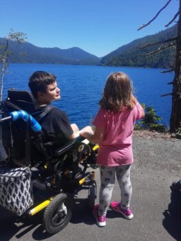 A teen boy in a wheelchair looks at a little girl standing next to him while overlooking a lake