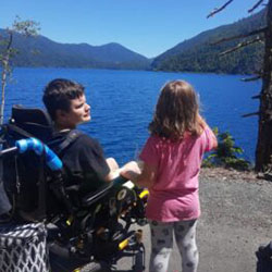 A teen boy in a wheelchair looks at a little girl standing next to him while overlooking a lake