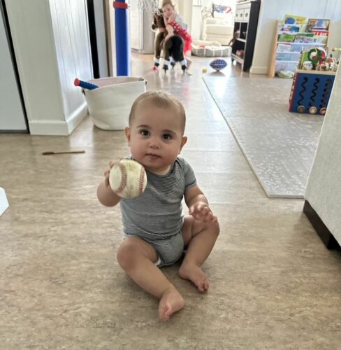 an infant holding a baseball up to the camera while sitting on the floor