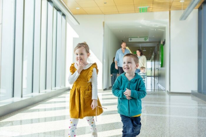 A young brother and sister smile as they walk across a skybridge at Seattle Children's Hospital