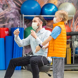 A child life therapist holds a video game controller while a young boy watches, patiently waiting for his turn to play