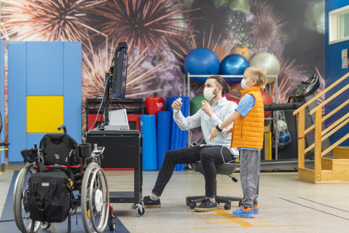 A child life therapist holds a video game controller while a young boy watches, patiently waiting for his turn to play