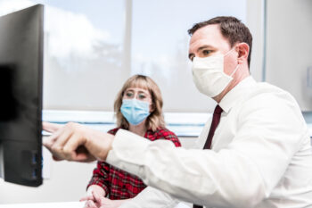 two adults wearing masks, look at a computer screen