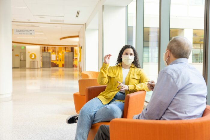A young woman and man wearing masks sit in orange chairs and chat