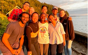 A group of adults participating in the Health Equity Track happily huddle together on a rocky beach with trees behind them while the golden glow of a sunset washes over them
