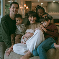 A family in neutral colored clothing poses and smiles at the camera in a living room setting.