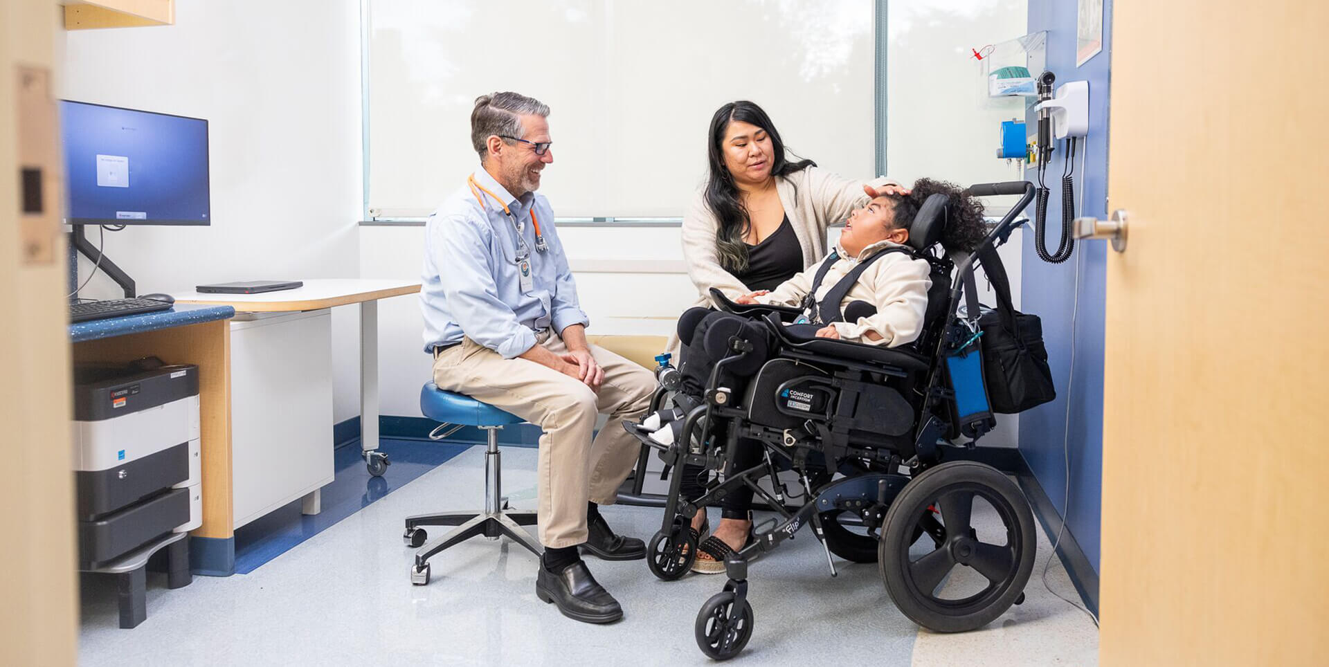A smiling pediatrician interacting with a young patient using a wheelchair and their mother in a bright exam room.