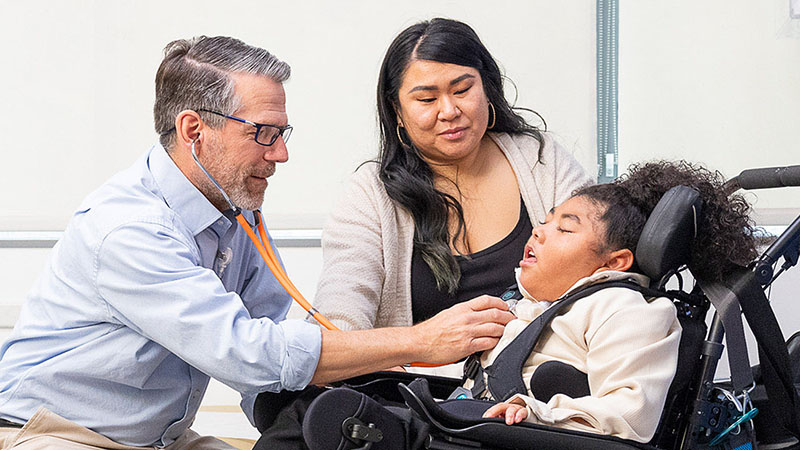 A doctor examines a patient in a wheelchair while the patient's mother looks on
