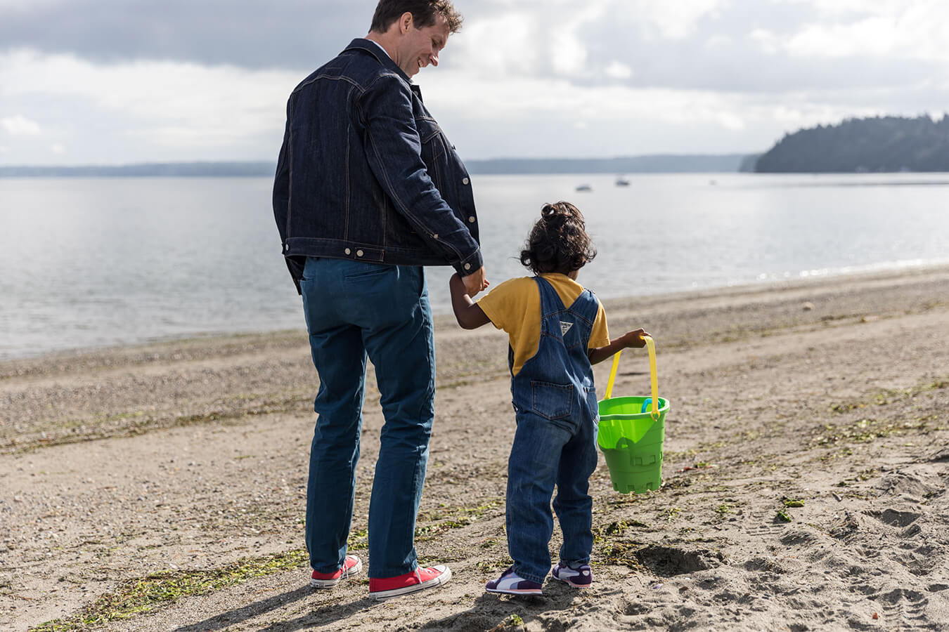 Father and child at beach. Child in yellow shirt carries a green toy bucket