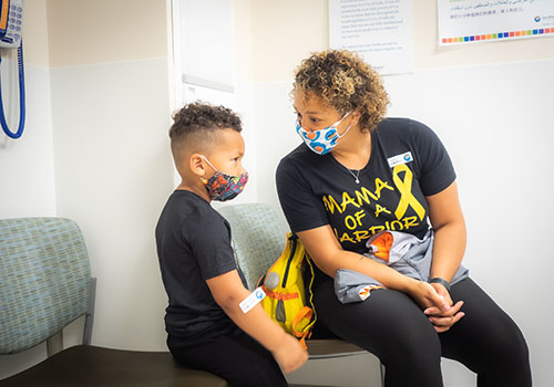 Kelli (right) and her son (left) sit in a medical office and are talking. They are both wearing masks, and Kelli is wearing a shirt that says “Mama of a Warrior” with the yellow cancer awareness ribbon.