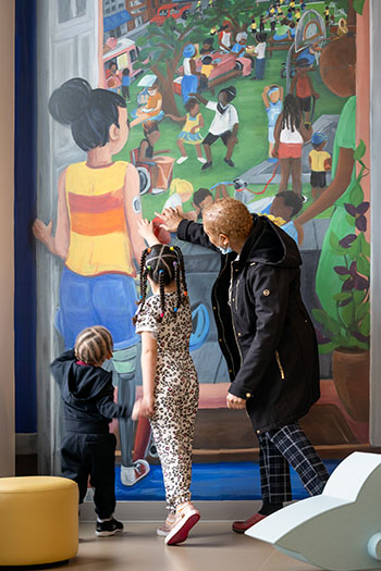 An adult and two children use their hands to feel a muraled wall at the OBCC Othello location. The mural is colorful and shows a diverse community of people interacting in a park.