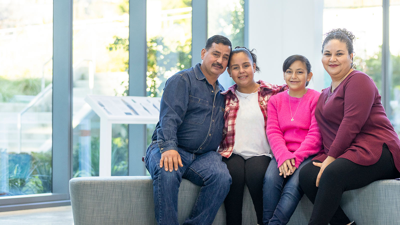 The Medina-Lamas family sits together on a gray bench at Seattle Children’s Hospital and smile at the camera. Parents Martin and Ana sit on either sides of their two daughters Mary and Monica.
