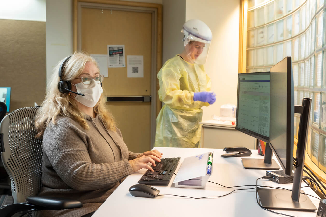 In the foreground, a nurse works on a computer in Seattle Children’s Pre-Admission Testing Coordination clinic. In the background, a nurse in protective gear.