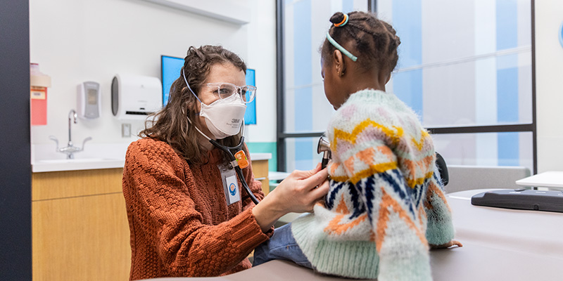 A female doctor with a mask on uses a stethoscope to listen to a little girl's heart