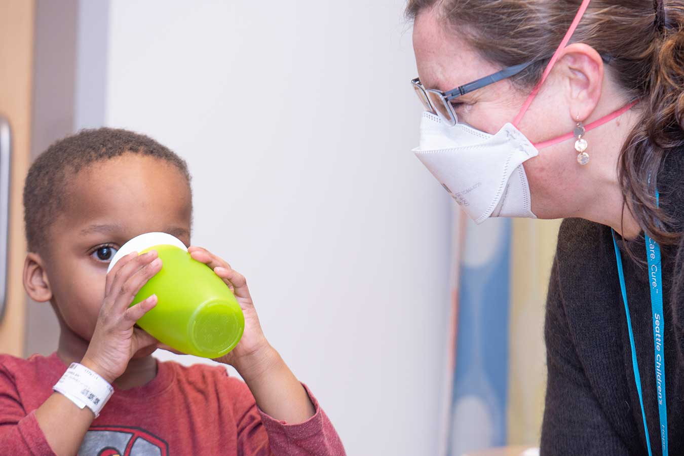 A young boy in a red top drinks from a green cup with a woman in a mask and glasses looking on.