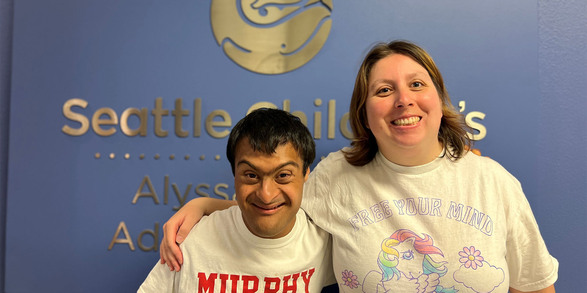 A male student and his caregiver stand in front of the Seattle Children's Alyssa Burnett Adult Life Center sign on a blue wall inside the center