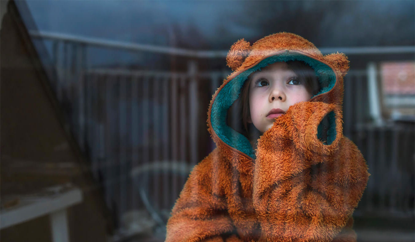 A child in a bear hoodie sits looking out a window