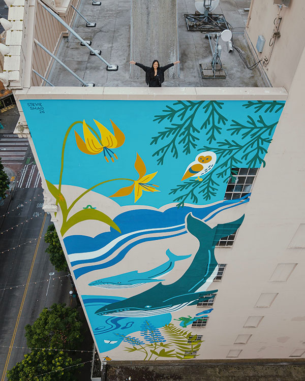 A woman stands on top of a building. The side of the building features her mural