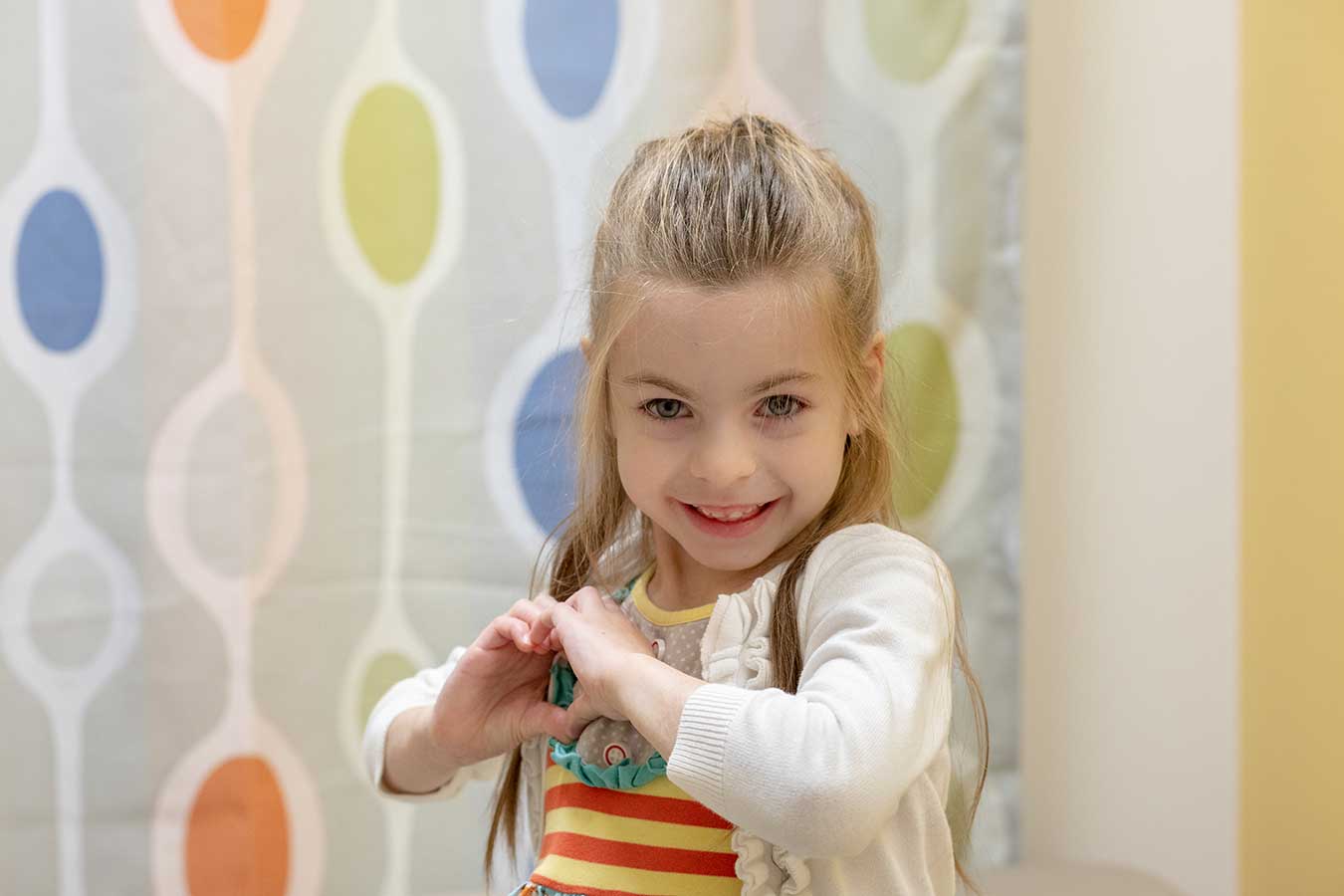 Young girl standing in front of colorful wall makes a heart shape with her hands
