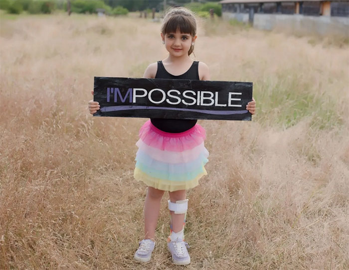A girl stands in a field holding a sign that reads "I'M POSSIBLE"
