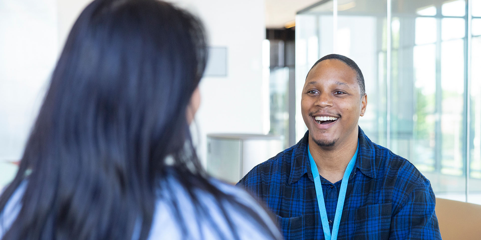 A Black man with short hair and a goatee, wearing a blue and black plaid shirt and a bright teal lanyard, laughing while looking at a woman with long dark hair whose back is to the camera in a bright, modern office.