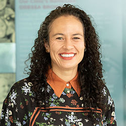 Shaquita Bell, MD, a woman with long, curly dark hair and a warm smile, looking at the camera against a soft teal background. She is wearing an orange collared shirt layered under a black blouse with a colorful floral and bird pattern.