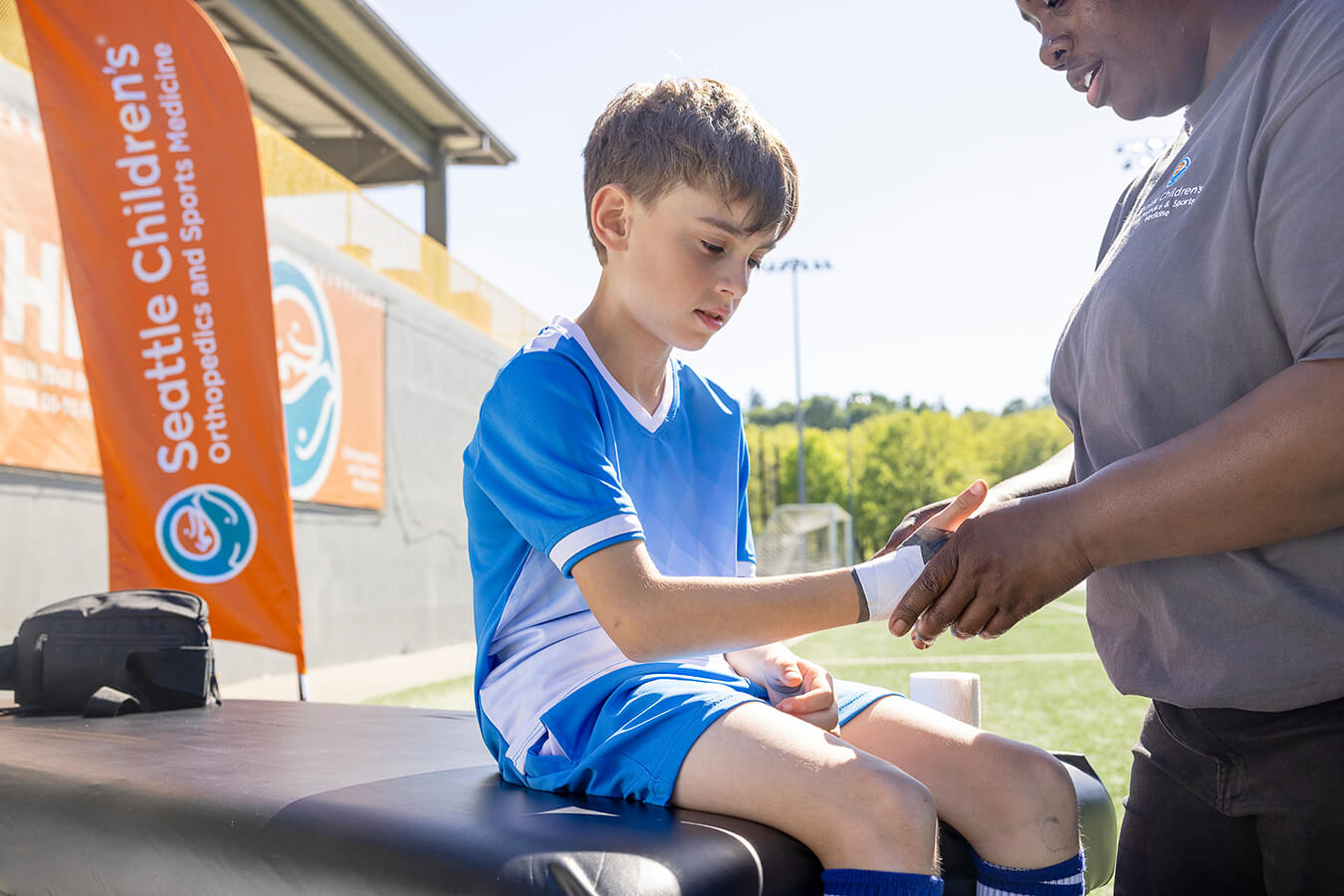 A Seattle Children's specialist tapes the wrist of a young soccer player sitting on a sideline treatment table.