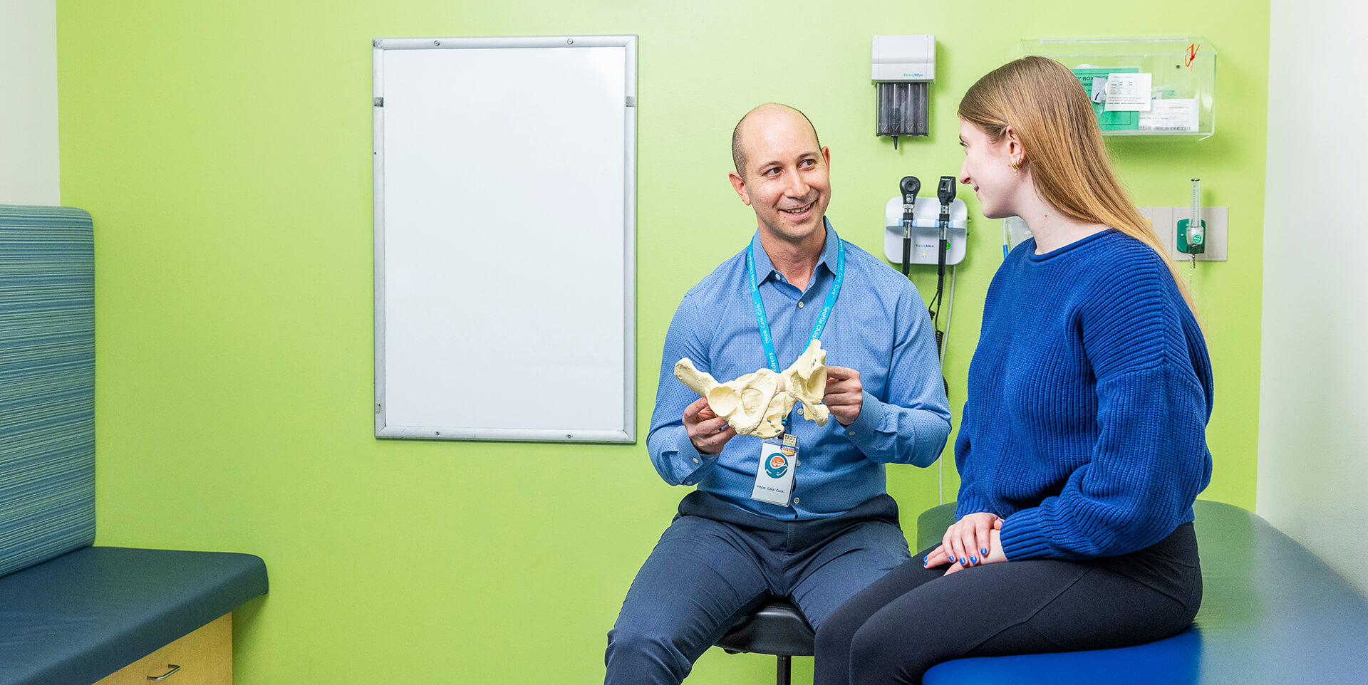 A Seattle Children's doctor uses a pelvic bone model to explain a hip condition to a teenage patient in an exam room.