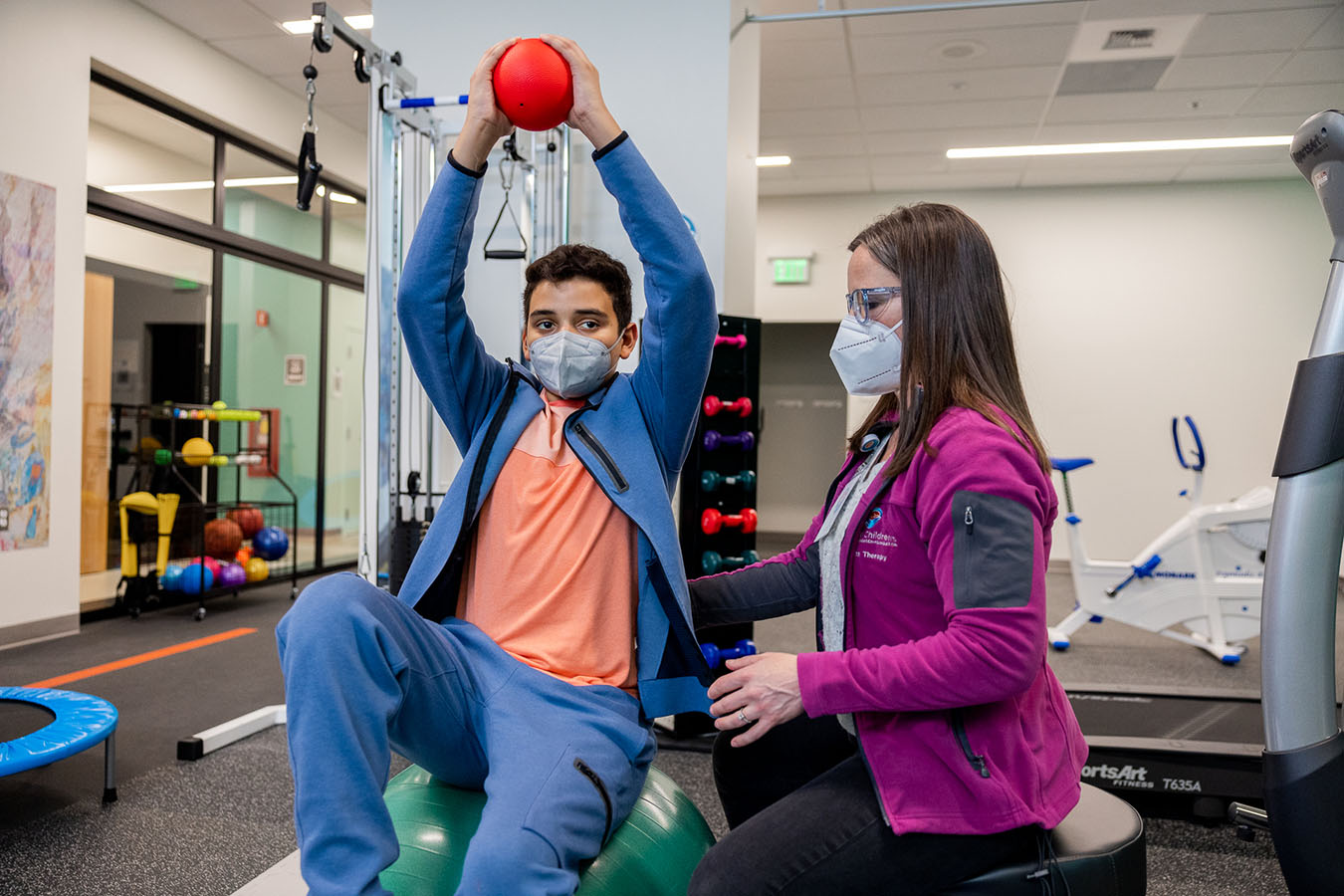 An adult guides a young man doing exercises in a gym