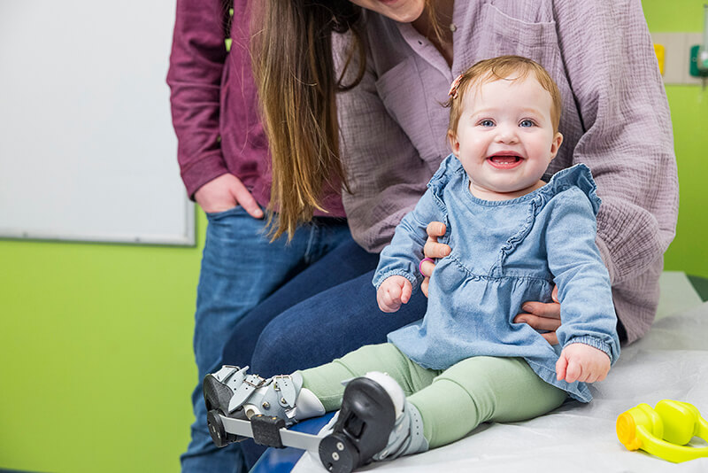 A smiling baby wearing a clubfoot brace with boots attached to a bar sits on an exam table while being supported by a caregiver.
