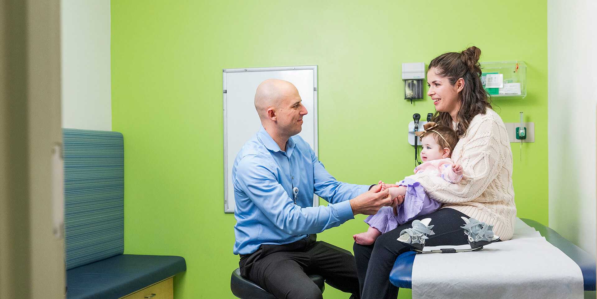 A Seattle Children's doctor examines a baby's foot as the baby sits on their mother's lap, with clubfoot bracing boots visible on the exam table nearby.