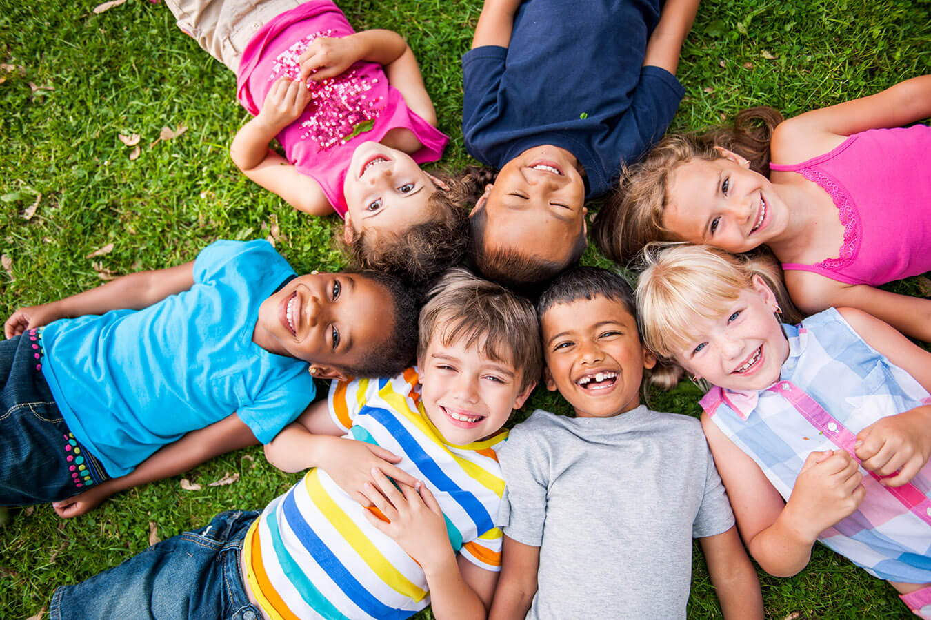 Seven diverse children lying in a circle on the grass and smiling upward.
