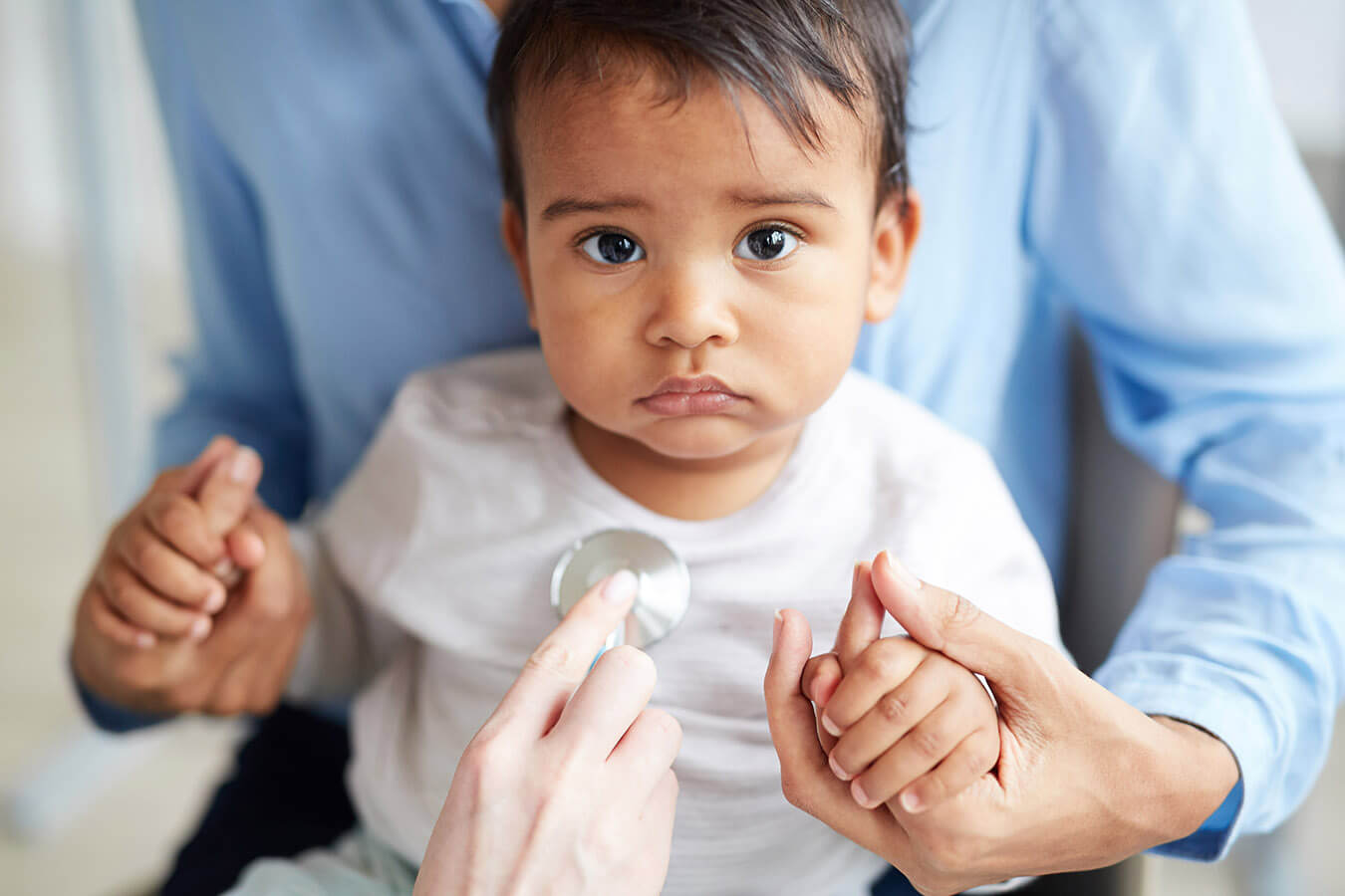 A healthcare provider using a stethoscope to examine a young child sitting in an adult's lap.