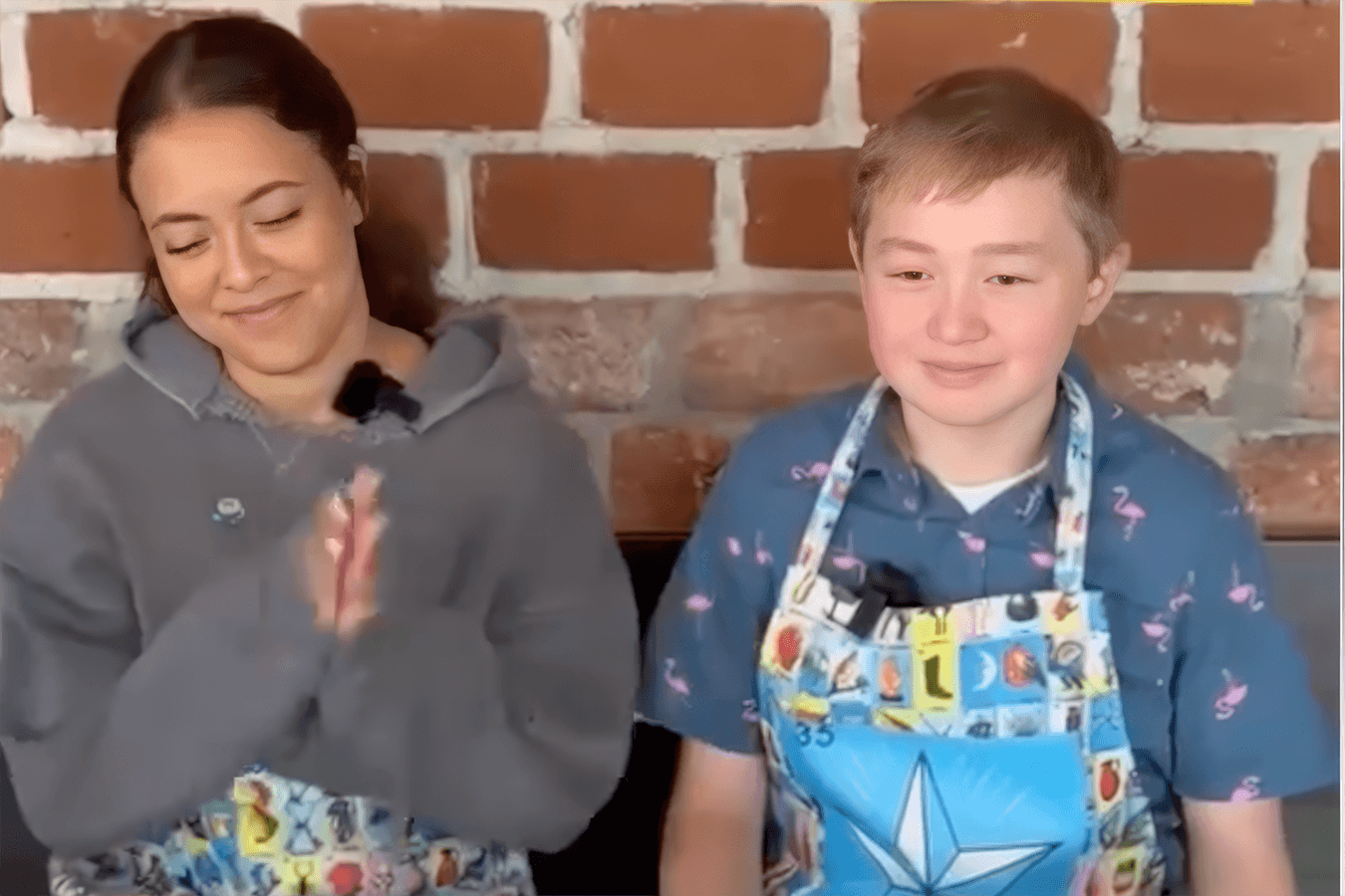 Two people wearing aprons sit in front of a brick wall, smiling and clapping.
