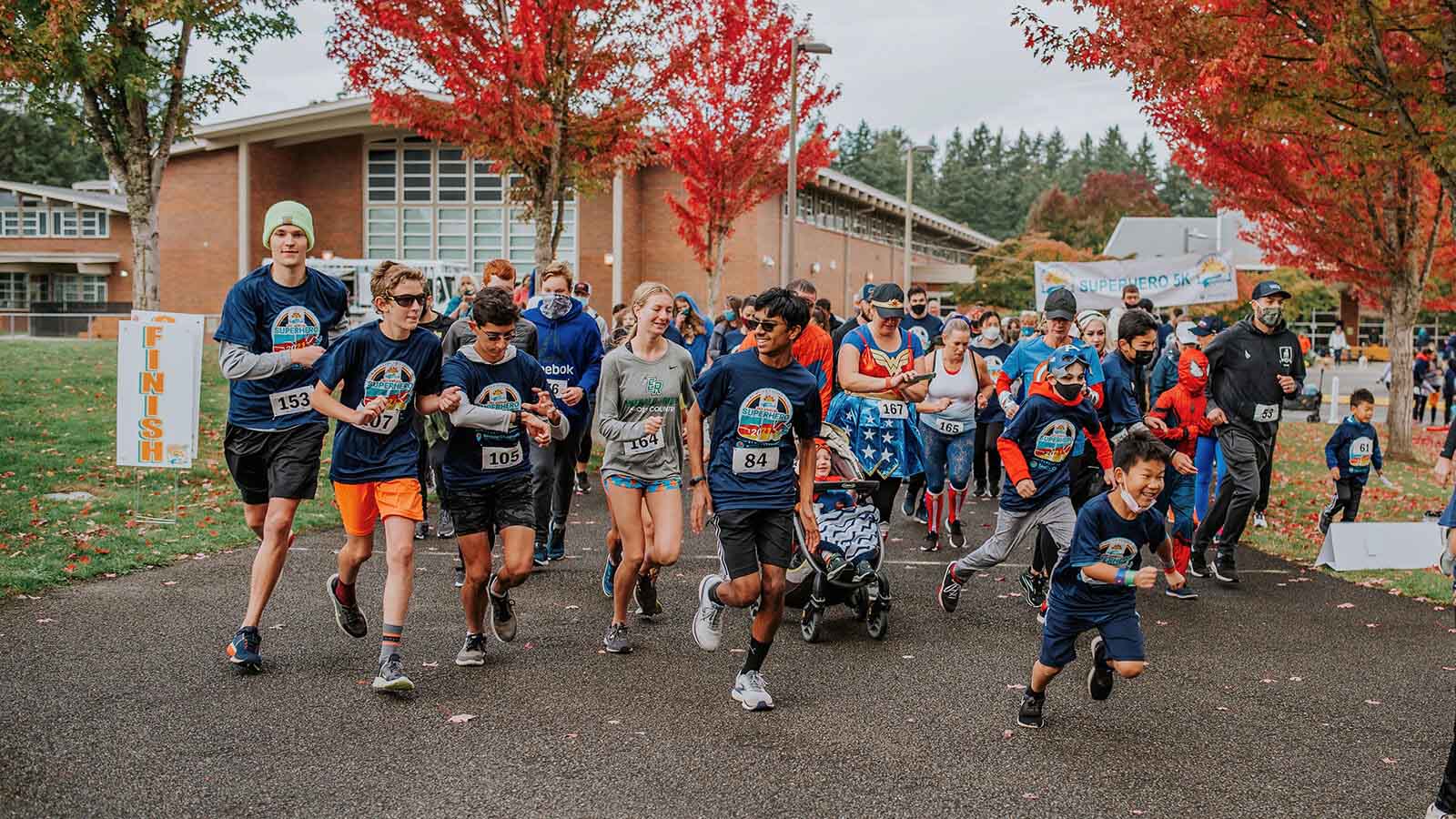 Runnners of all ages running from the starting line at the Superhero 5K surrounded by trees with red and orange fall leaves.